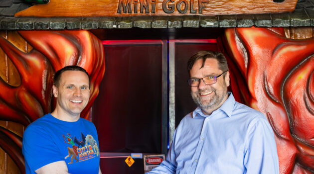 Two men stand outside of the enterance to Cryptid Mountain Miniature Golf shaking hands.