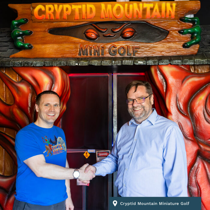 Two men stand outside of the enterance to Cryptid Mountain Miniature Golf shaking hands.